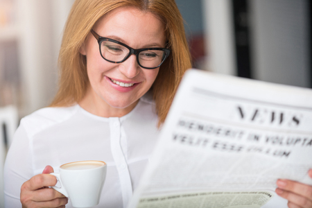Pleasant relaxation. Cheerful charming senior smiling woman drinking coffee and reading newspaper while sitting at the tableの写真素材