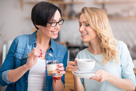 Yummy. Cheerful and smiling women eating sweet deserts and drinking coffee being in cafeの写真素材