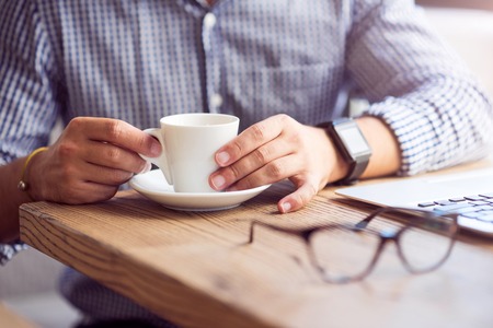 Favorite beverage. Close up of cup of coffee standing on the table while pleasant man drinking it and resting in the cafeの写真素材