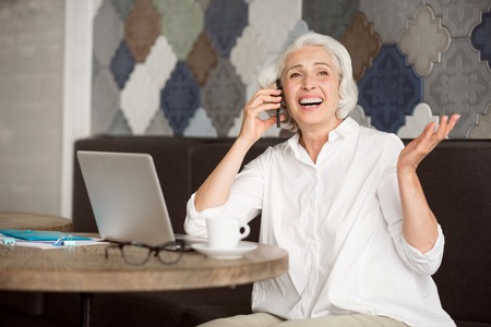 Live with inspiration. Positive delighted senior woman sitting at the table and smiling while talking on cell phoneの写真素材