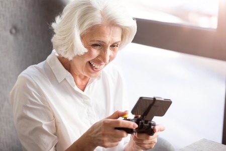 Like doing it. Overjoyed delighted senior woman sitting at the table and holding game console while playing video gamesの写真素材