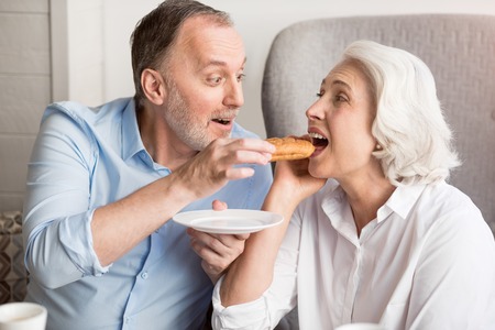 Lunch time. Positive delighted smiling husband feeding his wife with croissant and resting together in the cafeの写真素材