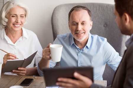 We like it. Positive delighted senior couple sitting at the table and talking with professional, sales manager while drinking teaの写真素材