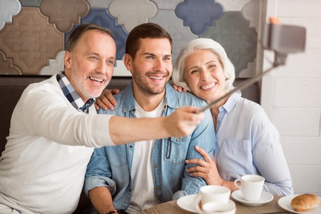 Happy together. Cheerful delighted smiling senior couple sitting at the table with their adult grandson and making selfies while resting in the cafeの写真素材