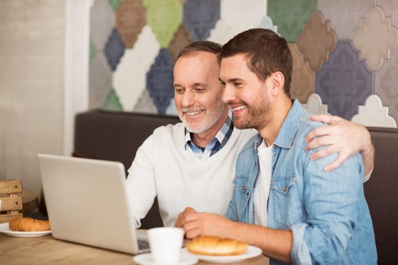 Full of positive emotions, Cheerful delighted smiling man and his grandson sitting at the table and using laptop while resting in the cafeの写真素材