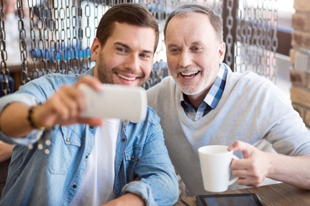 Remember this moment. Cheerful delighted man sitting at the table with his grandfather and resting while making selfiesの写真素材