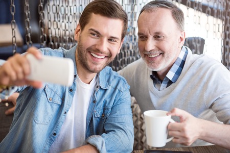Full of joy. Cheerful content smiling man and his grandfather sitting at the table and expressing positivity while making selfiesの写真素材