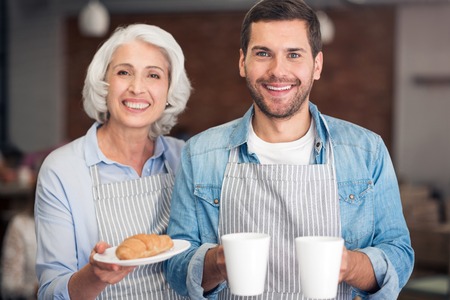 Hospitable people. Cheerful content woman and her adult grandson smiling and holding cup with tea and just baked croissantsの写真素材