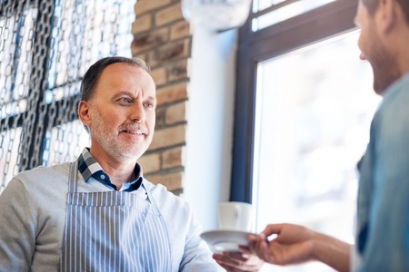 Here you are. Positive senior man holding cup of coffee and expressing gladness while talking with his colleagueの写真素材