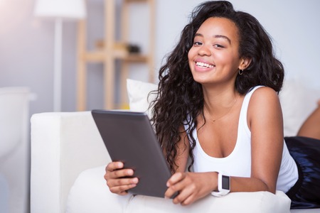 Enjoy relaxation. Cheerful beautiful smiling young woman lying on the couch and using tablet, while expressing positivityの写真素材
