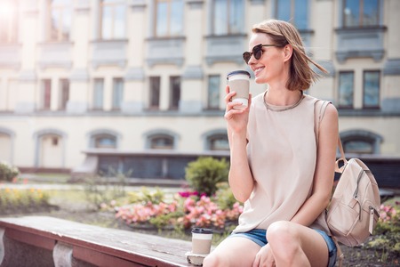 Good mood. Delighted and smiling young woman sitting and drinking coffee being outdoorsの写真素材