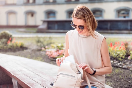 Stylish one. Positive young woman sitting and taking something out from her rose small backpackの写真素材