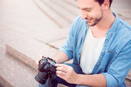 Like my work. Cropped image of smiling young man holding a photo camera sitting on steps outdoors