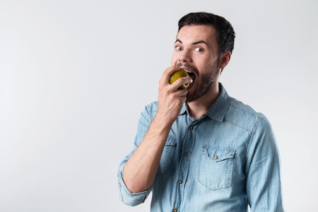 So tasty. Young handsome man staring and eating apple while standing against white background.