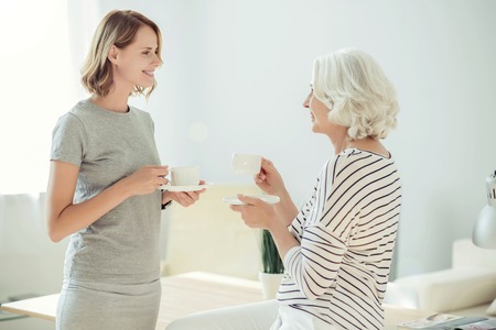 Pleasant relaxation. Cheerful delighted beautiful woman drinking coffee with her aged mother while resting together at homeの写真素材