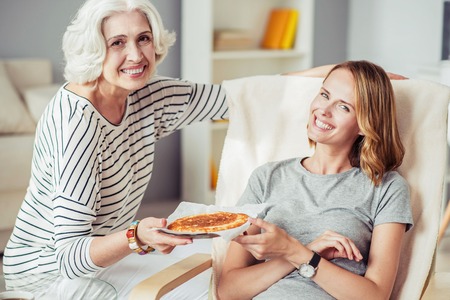 time for meal. Cheerful delighted smiling aged woman holding plate with pie and giving it to her adult daughter while resting together at homeの写真素材