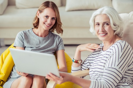 Spend time together. Cheerful smiling aged woman resting with her granddaughter and using laptop...