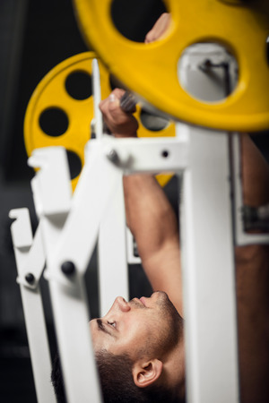 Exerting strength. Strong well built male weightlifter lying on a gym apparatus and looking at a barbell while pushing weightの写真素材