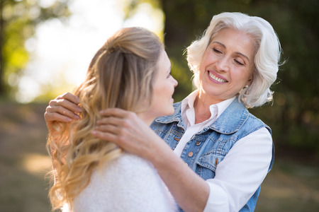 Soft care. Pretty grey haired woman smiling and playing with hair of her daughter while having rest in the park.の写真素材