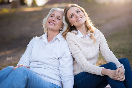 Dreams about future. Pleasant relaxed mother and daughter smiling and dreaming while sitting on the ground.の写真素材
