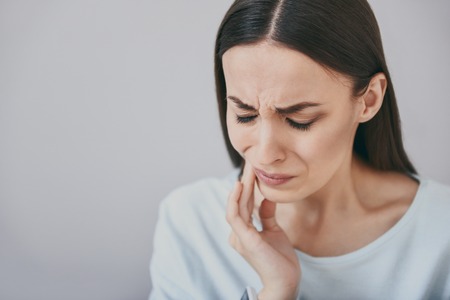 I need dentists help. Nervous young woman clothing her eyes and touching jowl while standing against isolated white background.の写真素材