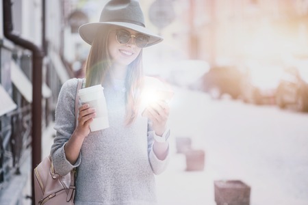 Have a nice time. Joyful delighted young woman using cell phone and typing messages while drinking coffee in the streetの写真素材