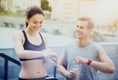 Here we go. Beautiful young athletic girl looking at her smartwatch while handsome man is holding a bottle of water while waiting for training.の写真素材