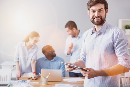 Working development. Young smiling bearded man standing on background of working colleagues with tablet in light office.の写真素材