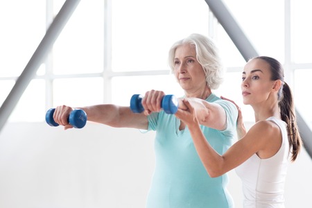 Develop your body. Serious confident persistent woman standing near the coach and looking forward while holding blue dumbbells in front of herの写真素材