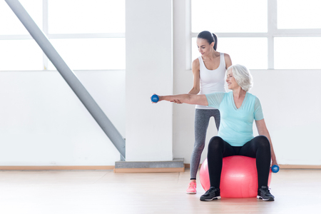 Various sports equipment. Pleasant confident slim woman holding blue rubber dumbbells and smiling while sitting on a fitness ballの写真素材