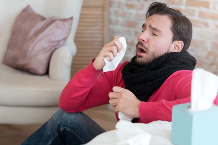 I hate being ill. Sick exhausted unhappy young man sitting on the floor and sneezing while holding a paper tissue in his handの写真素材