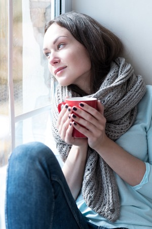 Looking outside. Nice relaxed dreamy woman sitting on a windowsill and having tea while looking...