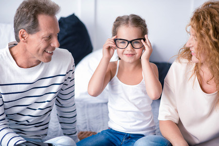 Look at my glasses. Cute little girl sitting on the bed with her grandparents while wearing glasses and having fun at homeの写真素材