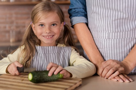 I can cook too. Joyful cheerful involved girl cutting a cucumber and sitting at the table in the kitchen next to her mother while cooking and having funの写真素材