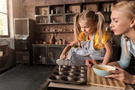 Involved in the cooking. Concentrated involved little girl putting the pastry in baking rings with her mother while sitting on the table in the kitchen and cookingの写真素材