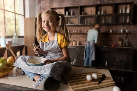 Enjoying the time in the kitchen. Nice delighted smiling girl holding the bowl and mixing eggs while sitting on the table in the kitchen and her mother cooking in the backgroundの写真素材