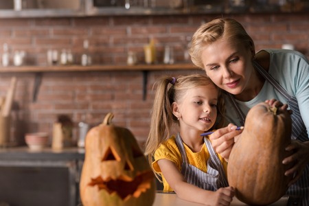 Lets do it together. Pleasant involved little girl drawing a face on the pumpkin for Halloween with her mother while standing in the kitchen near the table and expressing joyの写真素材