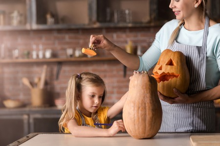We having fun. Amused funny smiling woman holding the pumpkin for Halloween and standing in the kitchen near the table while her daughter drawing a face on the pumpkinの写真素材