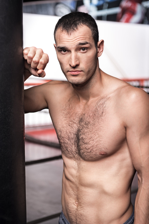 Amateur boxer. Handsome confident man standing near punching bag in the gym and leaning on it.の写真素材