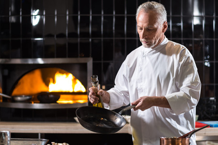 I enjoy the process of cooking. Concentrated involved aged chef holding the frying pan and the bottle of oil while standing near the table in the kitchen of a restaurant and cooking dishesの写真素材