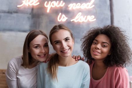 Happy together. Young cheerful delighted friends smiling and sitting close to each other while expressing joy at homeの写真素材