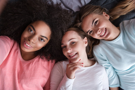 Joyful weekend. Cheerful delighted young girls smiling and lying on the bed while taking selfie and expressing joy in the bedroomの写真素材