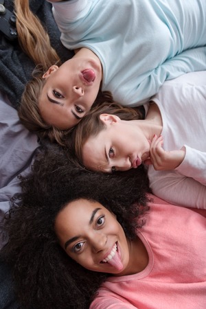 Funny selfie. Cheerful beautiful young girls smiling and lying on the bed while taking selfie and playing ape in the bedroomの写真素材