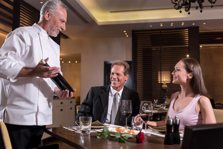 I recommend you this wine. Cheerfulsmiling bearded chef holding the bottle of wine while standing near the table in the restaurant and communicating with a coupleの写真素材