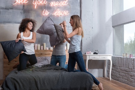 We having fun. Delighted amused young girls smiling and standing on the bed in the bedroom while having a funny pillow fightの写真素材