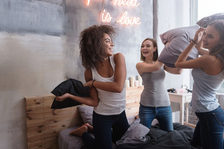 We playing funny games. Amused laughing young girls standing on the bed in the bedroom while having a funny pillow fight and expressing joyの写真素材