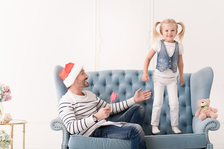 Having fun. Nice cute active girl standing on the sofa and jumping while having a great time with her fatherの写真素材