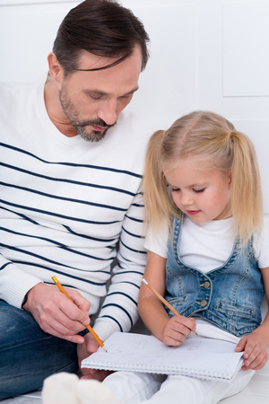 For better understanding. Handsome attractive intelligent man holding a pencil and looking at the copybook while explaining something to his daughterの写真素材