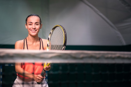 Play with me. Positive charming smiling young woman holding racket and playing tennis while training t in indoor courtの写真素材