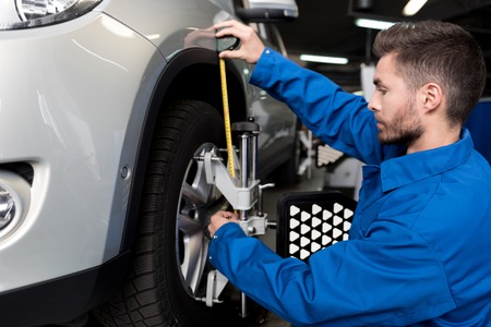 Pleasant young mechanic using measure tape and adjusting automobile wheel alignment while working in car stationの写真素材